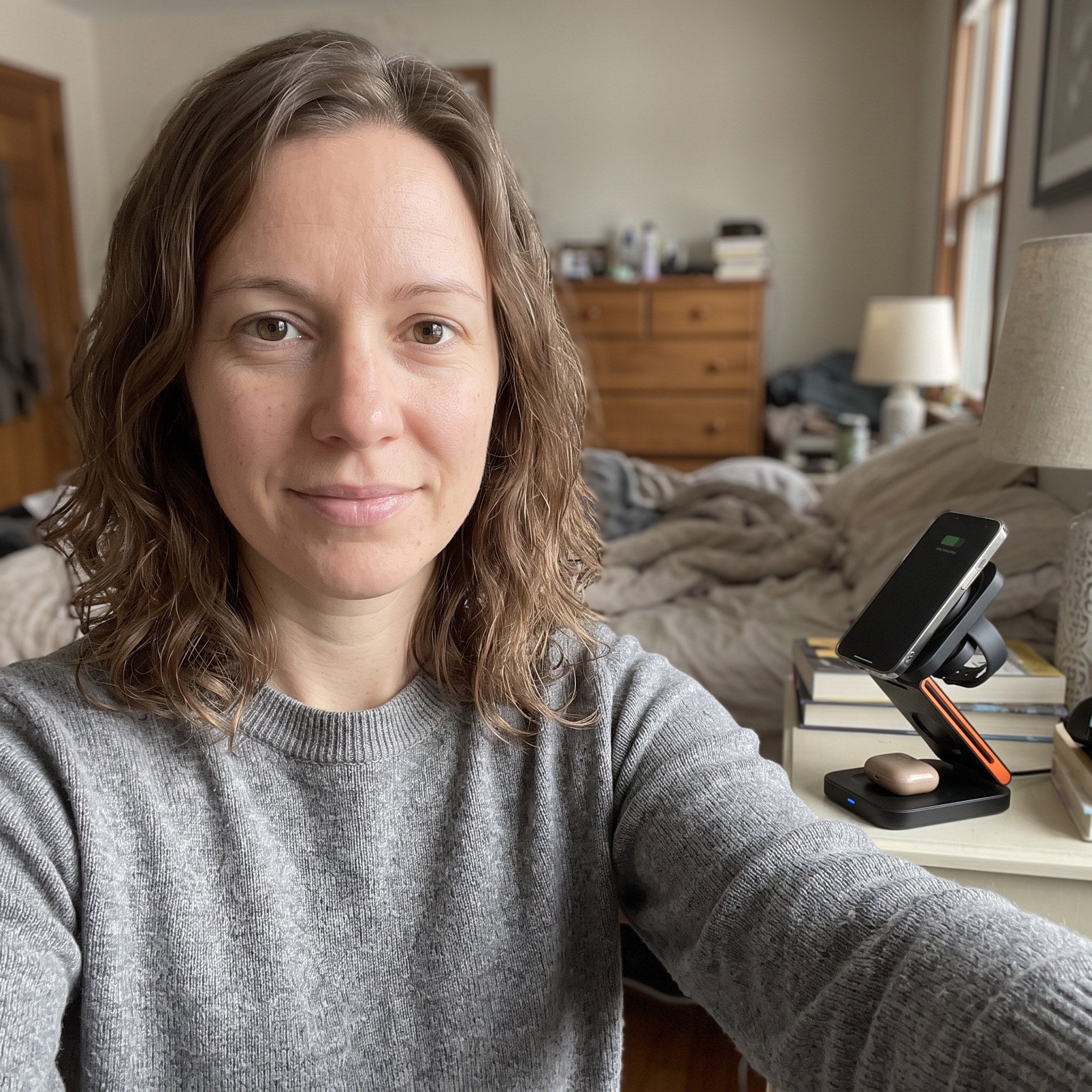 Self-shot adult woman at bedside, devices arranged on charger, informal lived-in room